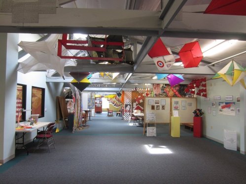A spacious indoor area with colorful kites and decorations hanging from the ceiling, featuring tables and art displays.