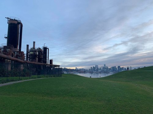 Industrial structures on the left with green hills and a city skyline across the water under a cloudy sky.
