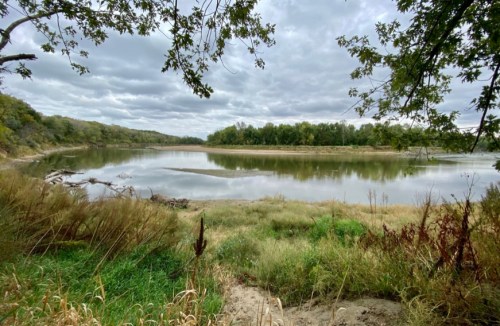 A serene river scene surrounded by lush greenery and cloudy skies, reflecting the landscape in calm waters.
