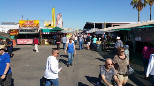 A bustling outdoor market with vendors, colorful banners, and people walking along the pathways under a clear blue sky.