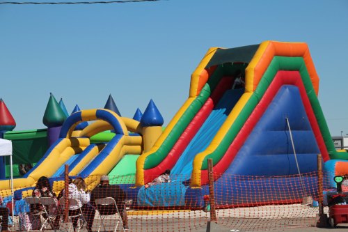 Colorful inflatable slides and bounce houses at an outdoor event, with people sitting nearby.