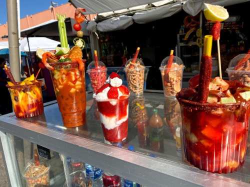 A colorful display of various drinks and snacks, including fruit cups and cocktails, at a vibrant outdoor market.