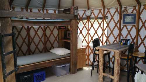 Interior of a cozy yurt featuring a bunk bed, wooden table, and rustic decor. Natural light filters through the windows.