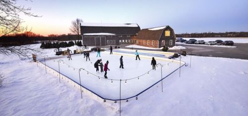 Outdoor ice skating rink surrounded by snow, with people skating and a barn in the background at sunset.