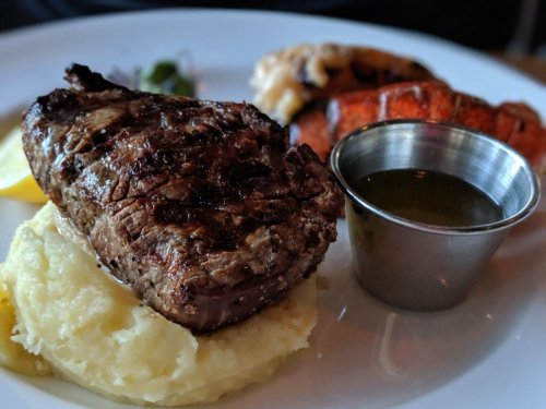A plate featuring a grilled steak on mashed potatoes, with lobster and a small cup of sauce on the side.