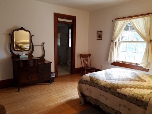 A cozy bedroom featuring a bed with a quilt, a wooden dresser, a rocking chair, and a window with curtains.