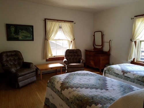 A cozy bedroom with two beds, a wooden table, armchairs, and large windows with light curtains.