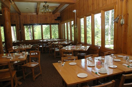 A cozy wooden dining room with empty tables set for a meal, large windows showcasing a green outdoor view.