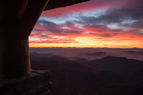 A vibrant sunrise over misty mountains, viewed from a rustic wooden structure.