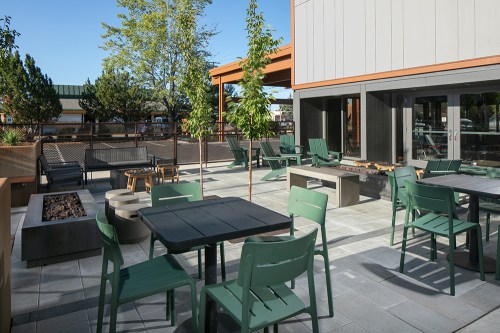Outdoor patio area with green chairs, tables, and fire pits surrounded by trees and modern architecture.