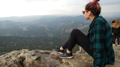 A person in a plaid shirt sits on a rocky ledge, gazing at a scenic mountain view.