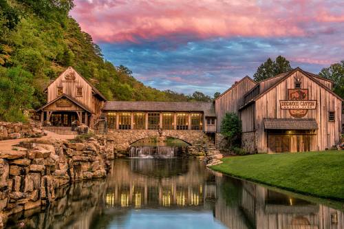 A rustic building by a serene pond, with a waterfall and colorful sunset sky in the background.