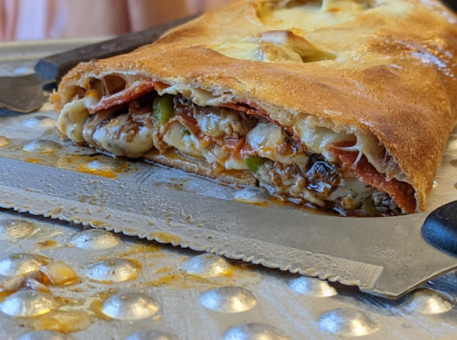 Close-up of a calzone filled with cheese, meats, and vegetables, resting on a metal surface with a knife beside it.