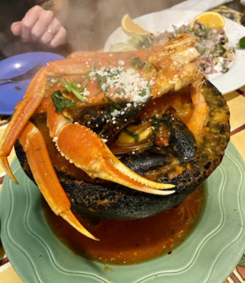A steaming seafood dish with crab, mussels, and herbs served in a black stone bowl, accompanied by a fresh salad.