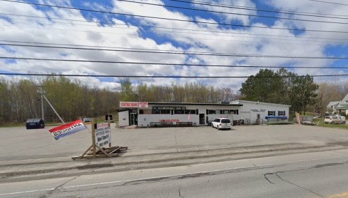 A roadside view of a small business with a sign, surrounded by trees and a cloudy sky. A car is parked in front.