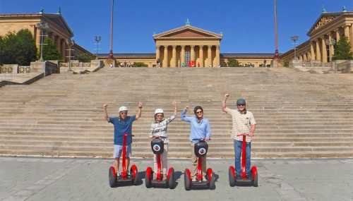 Four people on red segways wave in front of the Philadelphia Museum of Art, with steps and blue sky in the background.