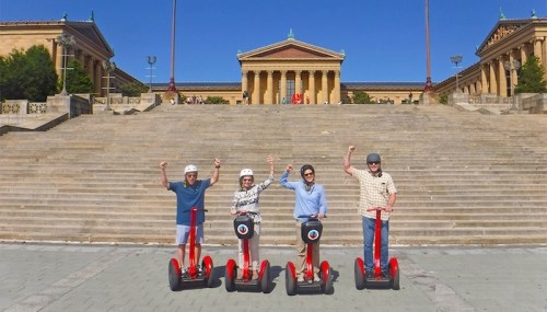 Four people on red segways wave in front of the Philadelphia Museum of Art, with steps and blue sky in the background.