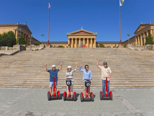 Four people on red Segways pose with raised arms in front of the Philadelphia Museum of Art on a sunny day.
