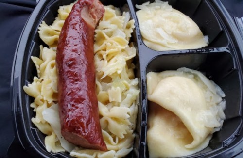 A divided meal tray featuring noodles with cabbage, a sausage, and two servings of dumplings.