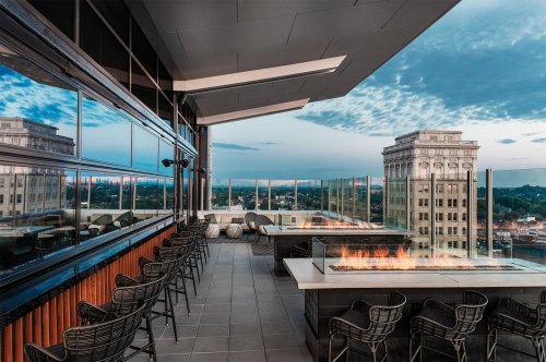 Rooftop lounge with modern seating, fire pit, and city skyline views under a blue sky with scattered clouds.