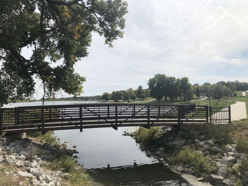 A wooden bridge spans a calm waterway, surrounded by greenery and a park in the background under a cloudy sky.