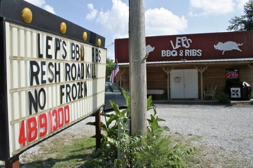 A BBQ restaurant with a sign reading "Fresh Roadkill, No Frozen" and an open flag, surrounded by gravel and greenery.