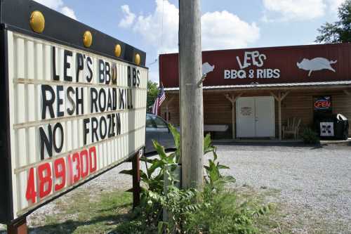 A BBQ restaurant with a sign reading "Fresh Roadkill, No Frozen" and an open flag, surrounded by gravel and greenery.