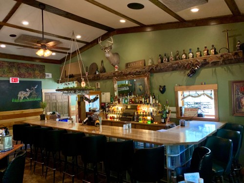 A cozy bar area with a wooden counter, shelves of liquor, and rustic decor, including a mounted deer head.