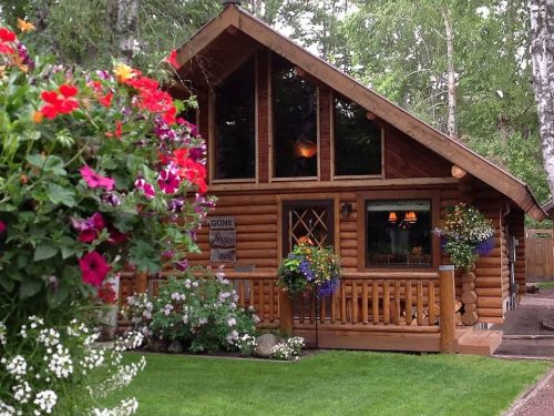 A charming log cabin surrounded by colorful flowers and greenery, featuring large windows and a welcoming porch.