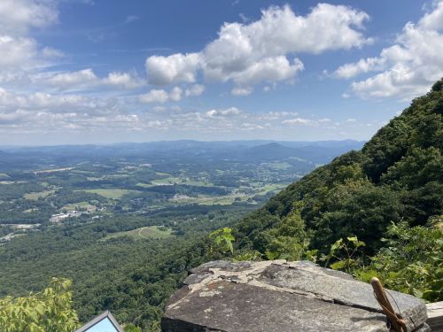 A scenic view from a mountain overlook, showcasing rolling hills and a blue sky with fluffy clouds.