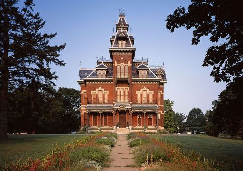 A grand Victorian mansion with intricate architecture, surrounded by lush greenery and colorful flowers.