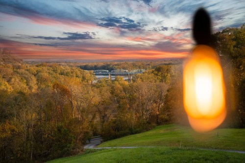 A sunset view over a river and bridge, framed by trees, with a blurred light bulb in the foreground.