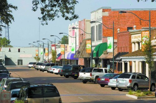 A quiet street lined with shops and parked cars, featuring colorful awnings and trees along the sidewalk.
