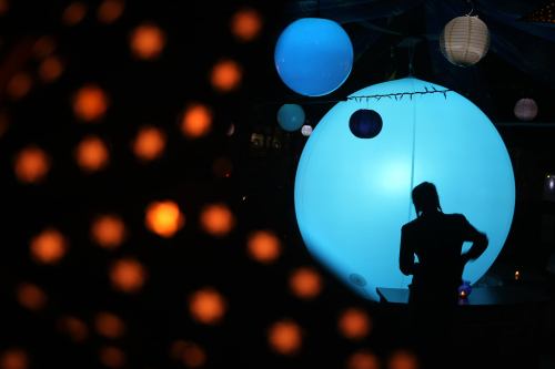 A silhouette of a person stands near a large blue sphere, surrounded by colorful lanterns and soft lighting.