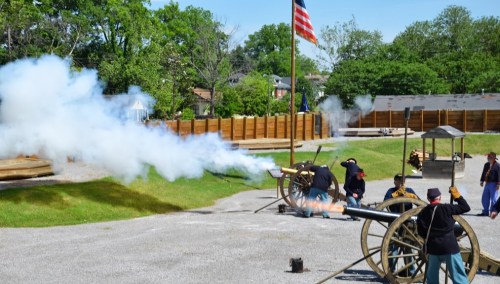 Civil War reenactors fire a cannon, with smoke billowing and an American flag waving in the background.