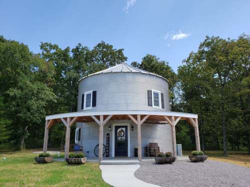 A round, silver metal building with a porch, surrounded by trees and a gravel path.