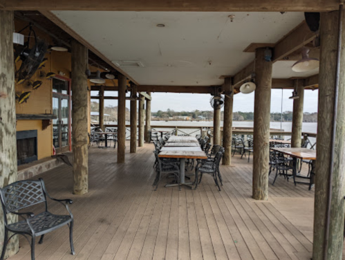 Outdoor dining area with wooden floors and tables, surrounded by pillars, overlooking a body of water.