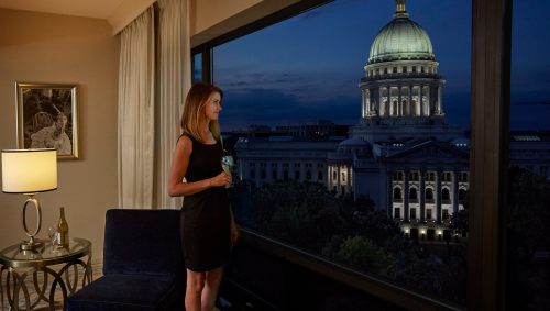 A woman in a black dress stands by a window, holding a drink, with a view of a dome building at night.
