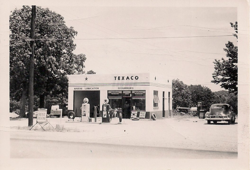 A vintage Texaco gas station with classic cars and retro signage, surrounded by trees and a clear sky.