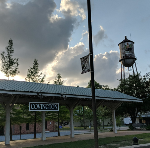 A sign reading "Covington" in front of a pavilion, with a water tower and cloudy sky in the background.