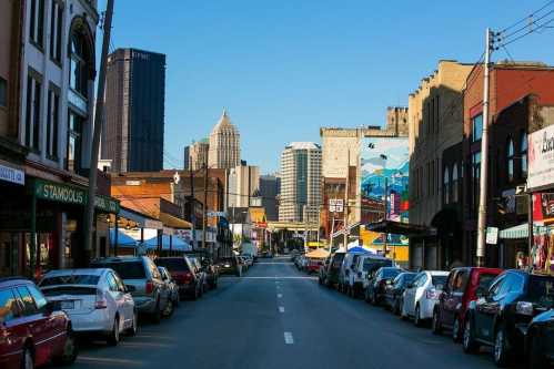 A city street lined with parked cars, leading to a skyline with tall buildings and colorful murals on the buildings.