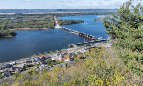 Aerial view of a river with a dam, surrounded by greenery and a small town along the shore.
