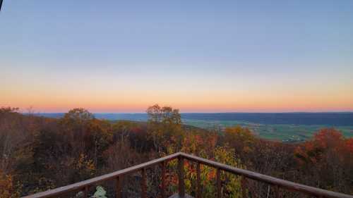 A scenic view from a wooden deck overlooking a valley with autumn foliage and a colorful sunset sky.