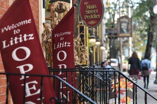 Two red banners reading "Welcome to Lititz" and "OPEN" outside a quaint shop, with autumn decorations and pedestrians nearby.