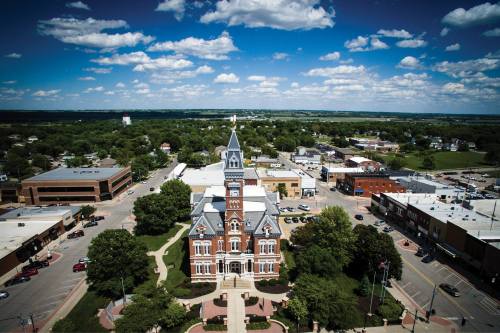 Aerial view of a town square featuring a historic courthouse surrounded by greenery and buildings under a blue sky.