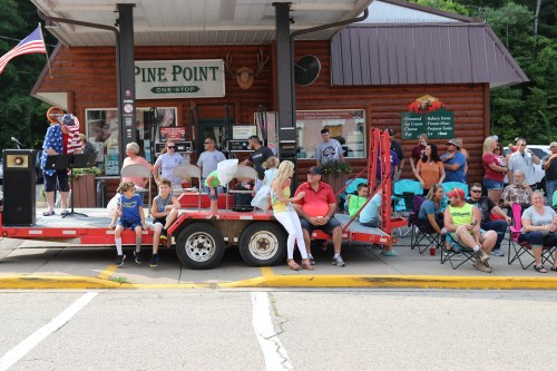 A crowd gathers at Pine Point for an outdoor event, with people seated on a trailer and a speaker on stage.