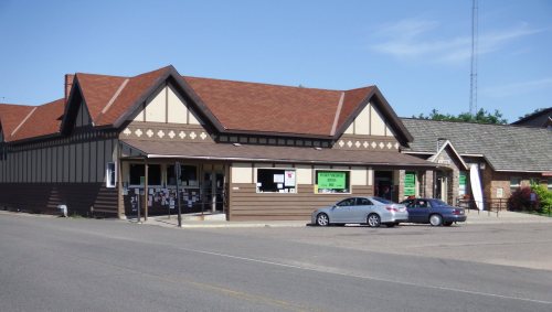 A two-story building with a brown exterior and a red roof, featuring large windows and signage, set on a quiet street.