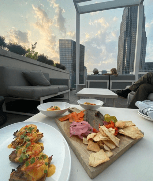 A rooftop dining scene with a platter of food and a city skyline at sunset in the background.