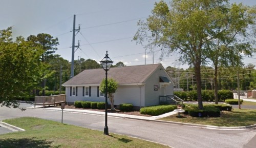 A small, light blue building with a sloped roof, surrounded by greenery and a lamp post, on a sunny day.
