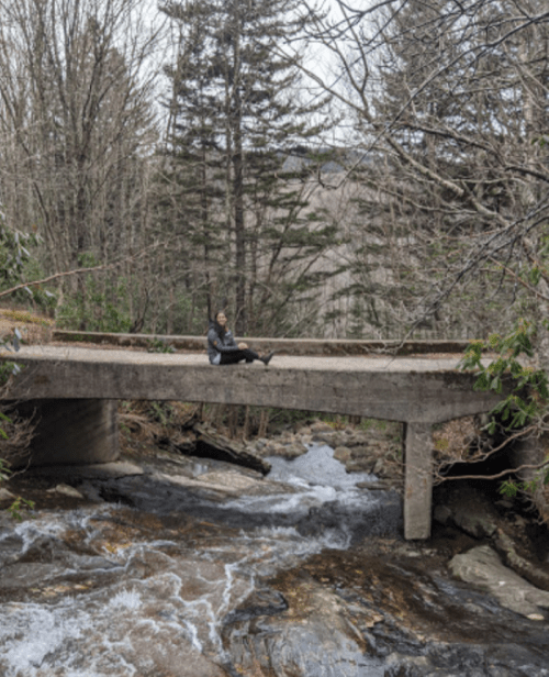 A person sits on a concrete bridge over a flowing stream, surrounded by trees and a rocky landscape.